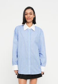 Žena v modro-bielej vertikálne pruhovanej blúzke s dlhými rukávmi, veľkým bielym golierom a manžetami, v kombinácii s čiernou plisovanou sukňou.