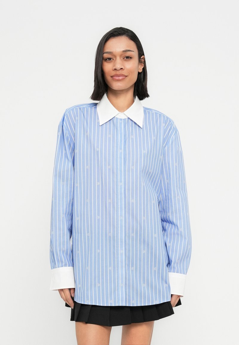 Žena v modro-bielej vertikálne pruhovanej blúzke s dlhými rukávmi, veľkým bielym golierom a manžetami, v kombinácii s čiernou plisovanou sukňou.