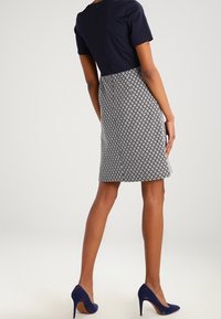 Kobieta ubrana w granatowy top, wzorzystą spódnicę do kolan i granatowe szpilki, stojąca na jasnym tle.