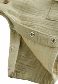 Tissu khaki clair avec une finition texturée, comportant des boutons-pression en métal et un bouton en bois. Comprend une poche et des détails de conception en couches.