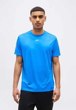Jovem vestindo uma t-shirt Reebok azul brilhante e calças pretas, em pé contra um fundo cinza claro liso, olhando para a frente.