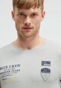 Graues Baumwoll-T-Shirt mit rundem Halsausschnitt, versehen mit blauer Druckschrift und zwei Stoff-Patches auf der Vorderseite. Der Text enthält "BLUE CREW" und "CAMP DAVID."