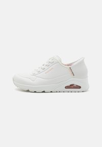 White sneaker with a textured upper, round toe, and lace closure. Features a cushioned sole with a visible air unit and subtle branding detail.