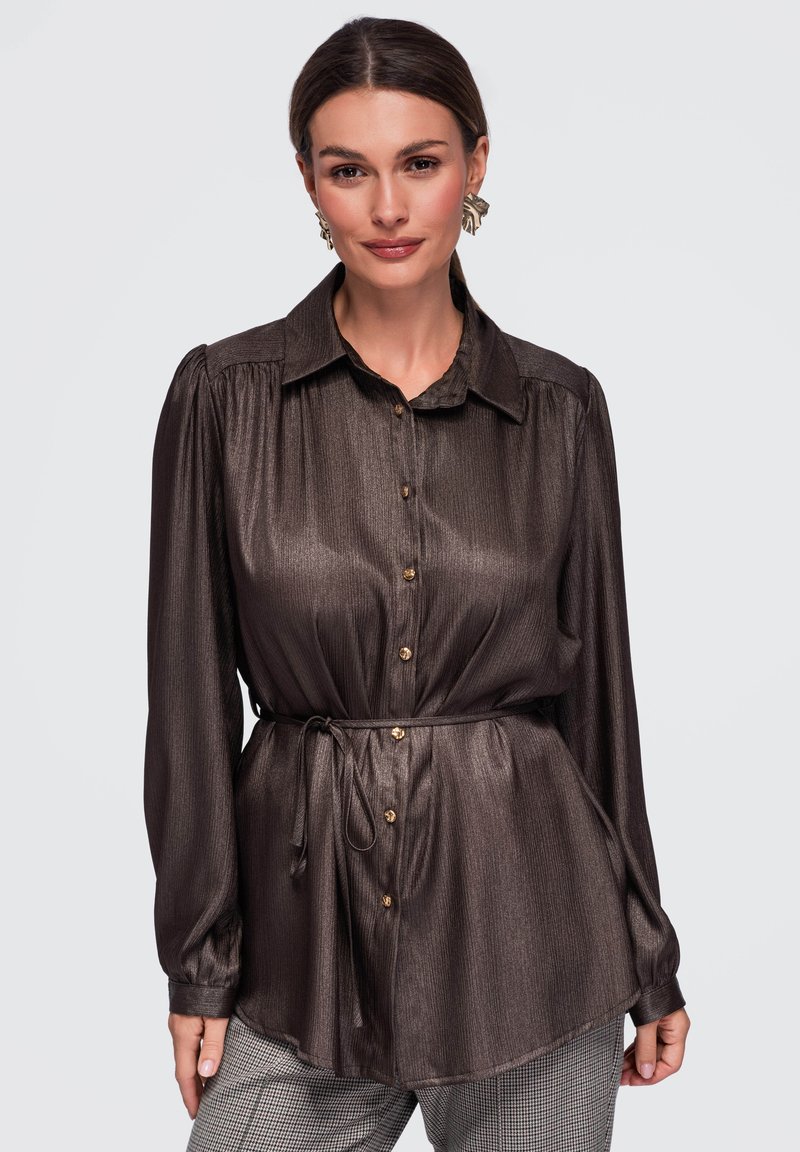 Femme portant un chemisier marron brillant à manches longues boutonné avec une ceinture à la taille et un pantalon à carreaux, posant devant un fond uni.