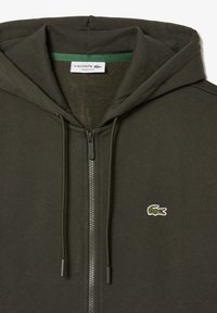 Haut à capuche zippé vert olive avec un tissu texturé, des cordons de serrage et un petit logo crocodile blanc sur la poitrine gauche. Design à coupe classique.