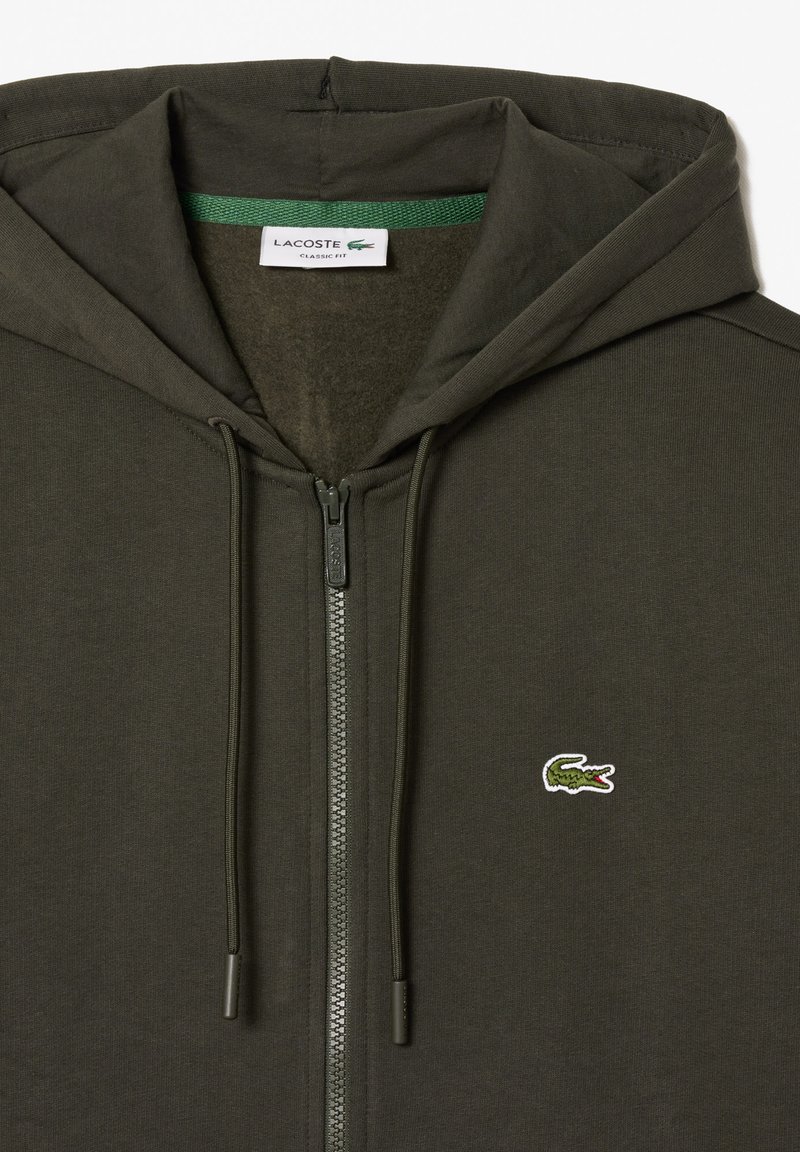 Olijfgroene zip-up hoodie met textuurstof, trekkoorden en een klein wit krokodillenlogo op de linkerkant van de borst. Klassieke pasvorm.