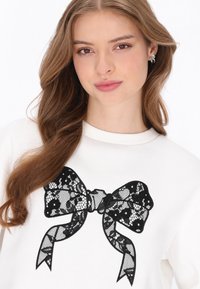 Fehér pulóver fekete csipkés masni mintával elöl, texturált kivitelben. A nyakkivágás kerek, és a szabás kényelmes, laza.