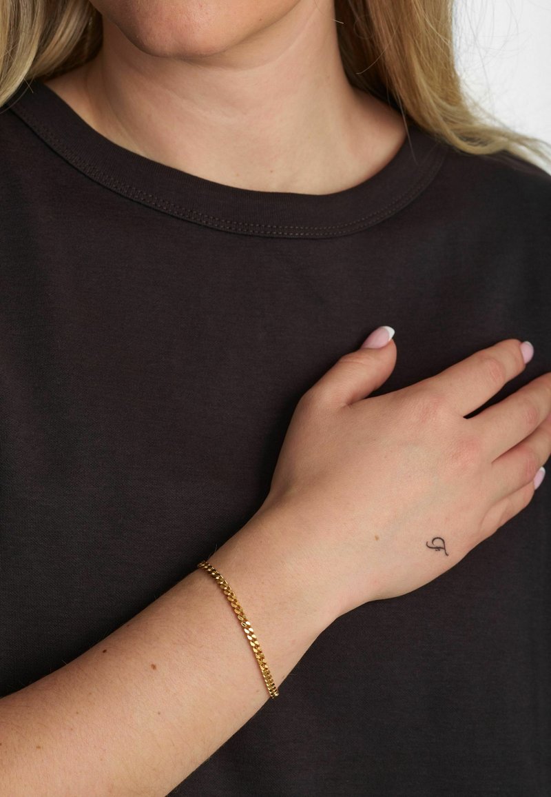 Braccialetto a catena d'oro su un polso, con un design testurizzato. Chi lo indossa ha una maglietta nera e un piccolo tatuaggio visibile sulla mano.