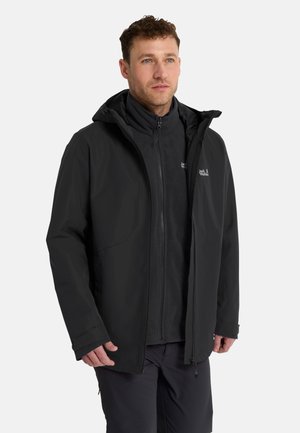 Man draagt een zwarte jas met rits en capuchon over een zwarte fleece, staand tegen een effen lichte achtergrond.