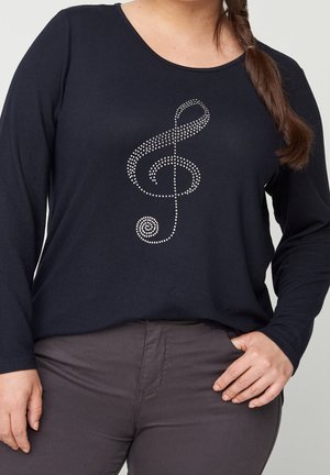 Femme portant un haut noir à manches longues avec un motif de clé de sol en strass et un pantalon foncé, debout avec une main dans la poche.