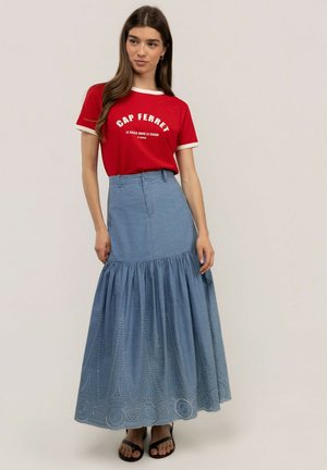 Joven mujer con cabello largo y castaño que lleva una camiseta roja de Cap Ferret, falda maxi azul con ojales y sandalias negras, de pie frente a un fondo liso.