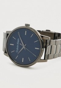 Ρολόι καρπού Calvin Klein με μπλε υφασμάτινο καντράν, ασημένιους δείκτες ωρών και βραχιόνι από ανοξείδωτο ατσάλι, εκτεθειμένο σε λευκό φόντο.