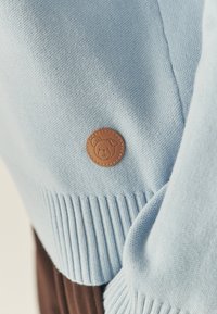 Hele sinine kootud sviiter ribitud mansettidega, millel on ümmargune beež nahkpatch, millel on karu nägu. Pehme tekstuur ja mugav disain.