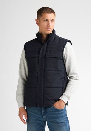 Mand iført navypolstret vest over lys grå sweatshirt og blå jeans, stående med den ene hånd i lommen, kiggende til siden.