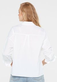 White shirt with long sleeves, relaxed fit, subtle sheen, and a back yoke detail. Fabric appears smooth, with rolled cuffs and no visible patterns.