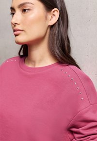Sweatshirt rose avec un col rond et des accents en strass le long de la couture de l'épaule. Fabriqué dans un tissu doux et lisse avec une coupe décontractée.