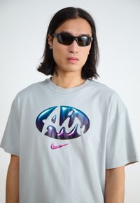 Világosszürke, túlméretezett póló, fényes, színes "Air" logóval és rózsaszín Nike pálcikával. A modellen fekete napszemüveg van.