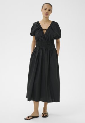 Femme portant une robe midi noire avec des manches ballon, une taille froncée, un col en V avec un lien, les mains dans les poches et des sandales plates noires.