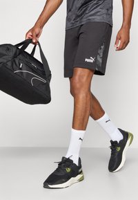 Calças de desporto pretas com um painel lateral de camuflagem, combinadas com ténis pretos e oliva, e meias brancas. Uma mala de desporto preta é segurada na mão.