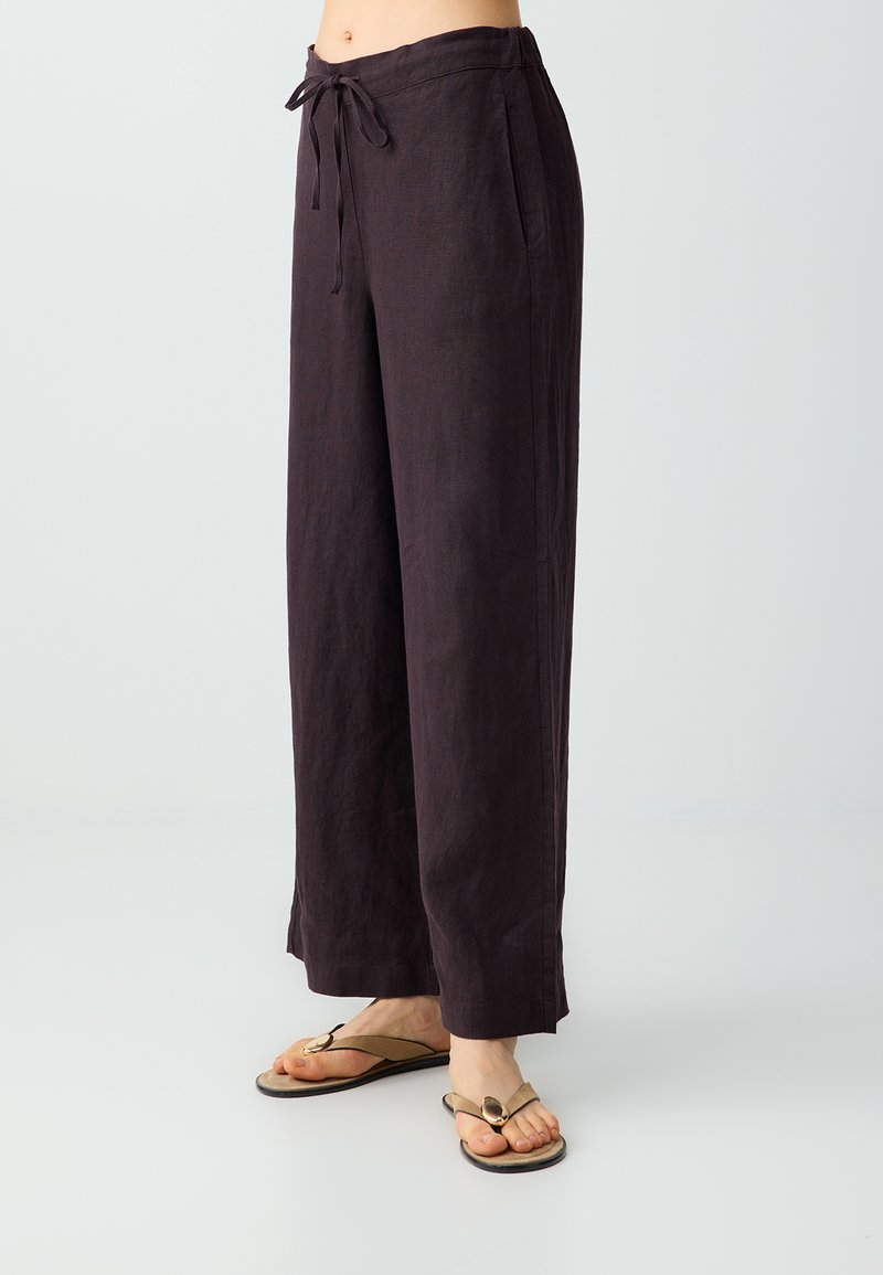 Personne portant un pantalon large marron foncé à taille coulissante et des sandales tongs beige, debout sur un fond clair.