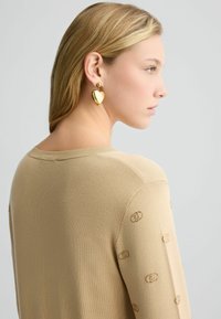 Maglione beige a maglia con scollatura rotonda, caratterizzato da delicati motivi a logo sulle maniche. Il modello indossa orecchini a forma di cuore in oro.