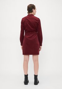 Burgundijas krāsas korduroja kleita ar apkakles dekoltē, garām piedurknēm un jostasvietu. Saskaņota ar melniem potīšu zābakiem. Gluda tekstūra.