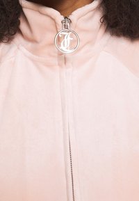 Ljusrosa jacka med dragkedja, tillverkad av mjukt, sammetslent material. Har en hög krage och en rund silvrig dragkedjeöppnare med logodetalj.