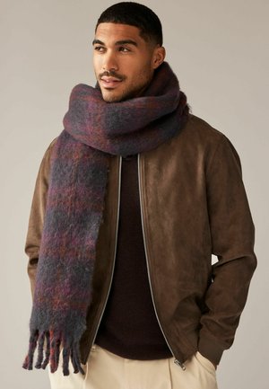 Wollschal in Grau-, Lila- und Rottönen, mit weicher Textur und fransigen Kanten, um den Hals eines Models gelegt, das eine braune Jacke trägt.