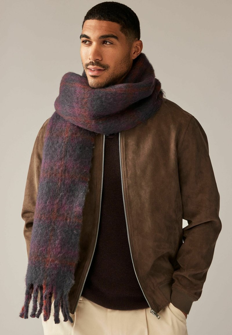 Wollschal in Grau-, Lila- und Rottönen, mit weicher Textur und fransigen Kanten, um den Hals eines Models gelegt, das eine braune Jacke trägt.