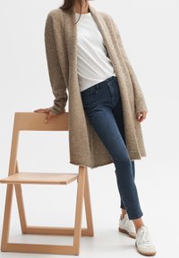 Femme appuyée sur une chaise en bois clair portant un long cardigan beige, un t-shirt blanc, un jean foncé et des baskets blanches sur un fond uni.