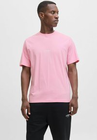 Camiseta de algodón rosa claro de manga corta con cuello redondo y un texto de logo blanco en el pecho, combinada con pantalones negros.