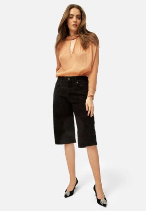Blusa pescozza con dettaglio a fiocco, abbinata a pantaloni corti neri in velluto a coste fino al ginocchio. La modella indossa tacchi neri con decorazioni.
