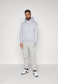 Lichtgrijze hoodie met kangoeroezak en geborduurd logo, gecombineerd met lichtgrijze joggingbroek en witte sneakers met donkere accenten.