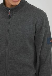 Gray zip-up jacket with a high collar, ribbed texture, and a logo patch on the left sleeve. Smooth fabric, casual design.