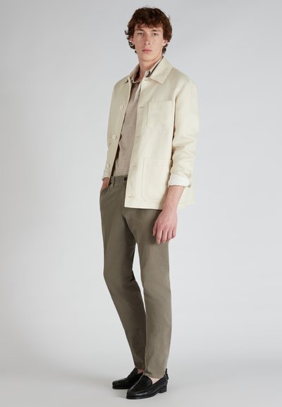 Giacca beige chiaro con taschino sul petto, abbinata a una camicia grigia e pantaloni verde oliva. Mocassini neri completano l'outfit.