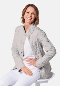 Hellgraue gesteppte Jacke mit Stehkragen, Druckknopfverschluss und strukturiertem Muster. Getragen über einem weißen Hemd, kombiniert mit weißen Hosen.