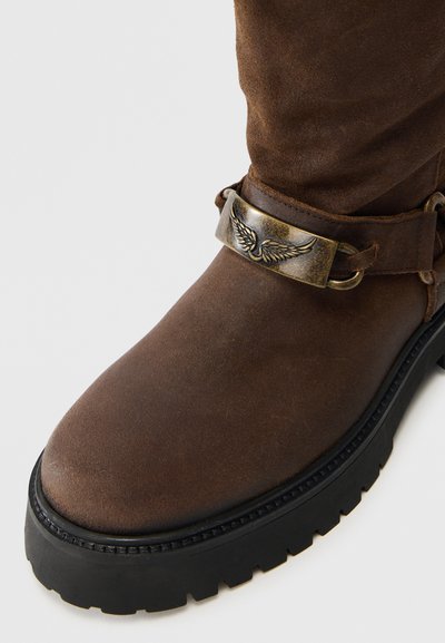 Καστόρινο καφέ μποτάκι που φτάνει στον αστράγαλο με στρογγυλεμένη μύτη, χοντρή μαύρη σόλα και μεταλλική ταινία με σχέδιο φτερού γύρω από τον αστράγαλο.