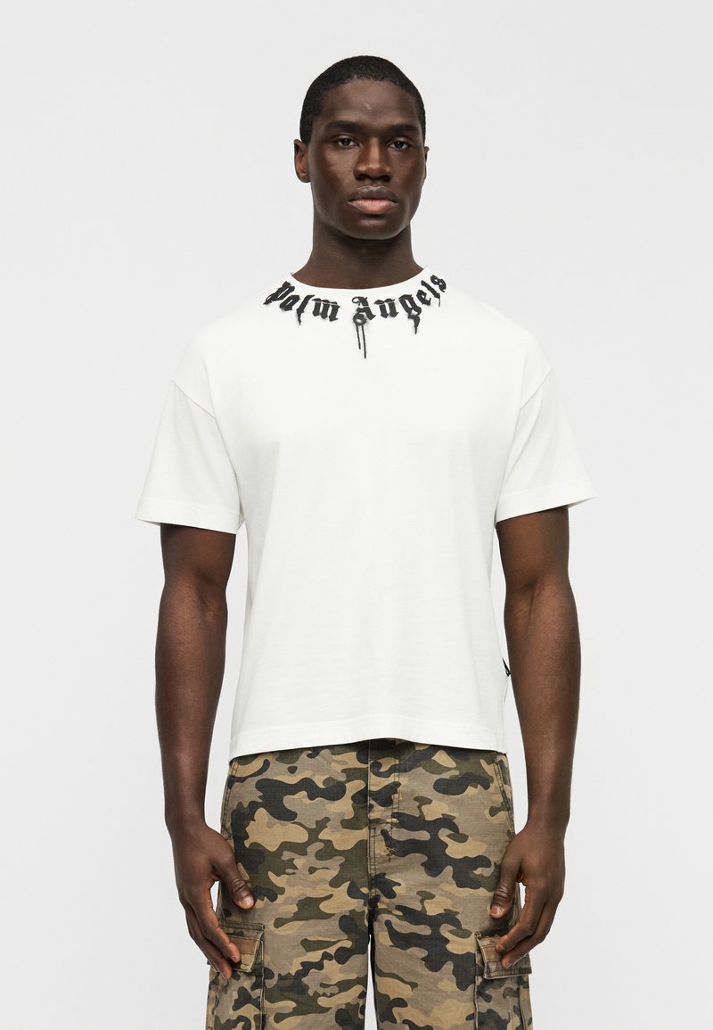Λευκό κοντομάνικο μπλουζάκι με μαύρο λογότυπο «Palm Angels» στον λαιμό, σε συνδυασμό με παραλλαγής παντελόνια cargo με πλαϊνές τσέπες.