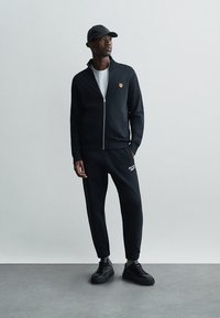 Black zip-up jacket with a small orange logo, paired with black joggers featuring white text. Worn with a black cap and black sneakers.