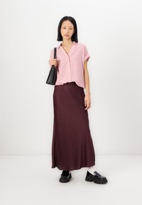 Gestreifte rosa Hemdbluse, fließender burgunderroter Satinrock und schwarze klobige Loafers. Das Model hält eine schwarze Handtasche und posiert vor einem weißen Hintergrund.