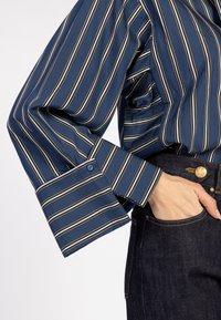 Blauw gestreept shirt met witte en lichtgrijze lijnen, voorzien van een brede manchette en een enkele blauwe knoop; gecombineerd met donkerdenim jeans.