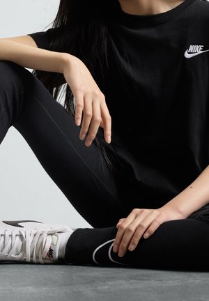 Osoba ubrana w czarną koszulkę Nike, czarne legginsy oraz białe buty sportowe Nike, siedząca na podłodze z jedną nogą zgiętą i rękami opartymi na nogach.