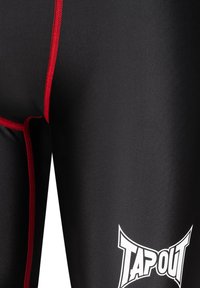 Melnas leggingas, kas izgatavotas no gluda materiāla, ar sarkanu šuvju detaļām un izteiktu baltu "TAPOUT" logo. Tuvplāns uz auduma tekstūru.