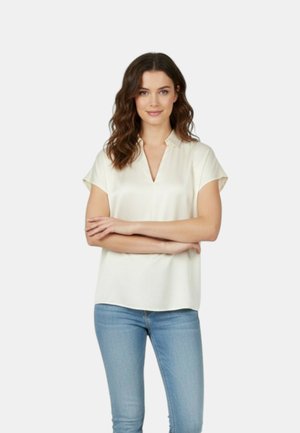 Giovane donna con capelli castani ondulati che indossa una blusa crema a maniche corte e jeans azzurri chiari, in piedi con le braccia incrociate e sorridente.