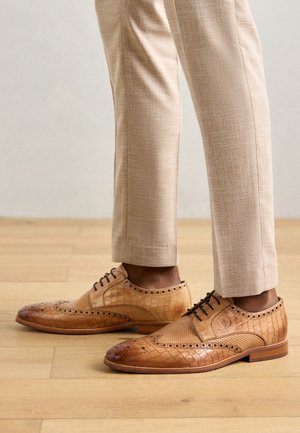 Helle Lederdressschuhe in Braun mit einem strukturierten Krokodilmuster, durchbrochenem Design und dunklen Schnürsenkeln, kombiniert mit hellbeigen Hosen.