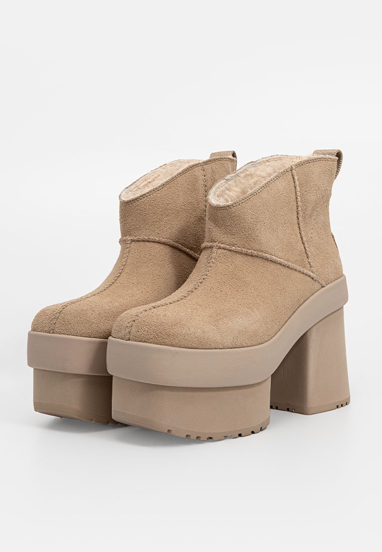 Un par de botas de tobillo de ante beige con plataforma, tacones gruesos y forro suave, exhibidas sobre un fondo blanco.
