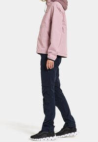 Personne debout portant une veste à capuche rose poussiéreux, un pantalon marine foncé avec poche zippée, et des baskets noires à semelles blanches sur fond blanc.