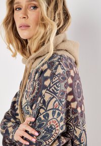 Sudadera con capucha estampada de base beige, con tejido texturizado, diseños florales coloridos en azul y burdeos, y una capucha suave y forrada de felpa.