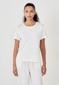 Weißes T-Shirt mit kurzen, aufgerollten Ärmeln, rundem Halsausschnitt und lockerer Passform. Weicher, glatter Stoff mit leichtem Glanz, kombiniert mit hellen Hosen.