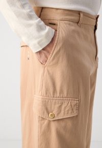 Beige cargo pants featuring a side pocket with a button closure, textured fabric, and visible stitching details.